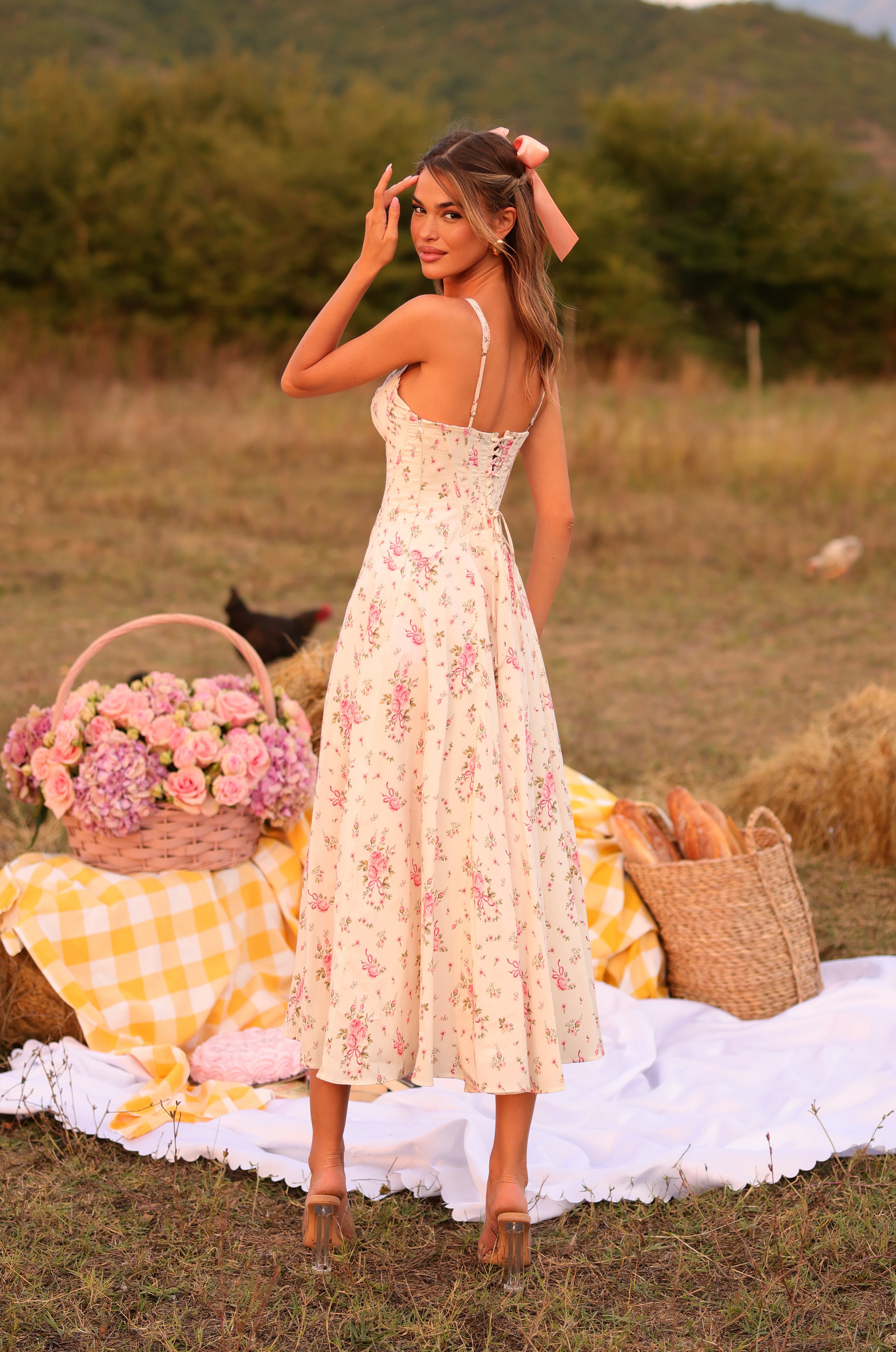 A woman wearing the Cream White Floral Casual Midi Dress: Rosella stands outdoors on a picnic blanket, with a basket of flowers and bread beside her and green hills in the background.